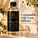 A bottle of MLA Marie Scents Luxe Gold fragrance oil stands on a stone surface with flowers and warm sunlight in the background. The bottle has a black cap and a black label with gold lettering, and the right side includes text reading 'Compatible with your diffuser.