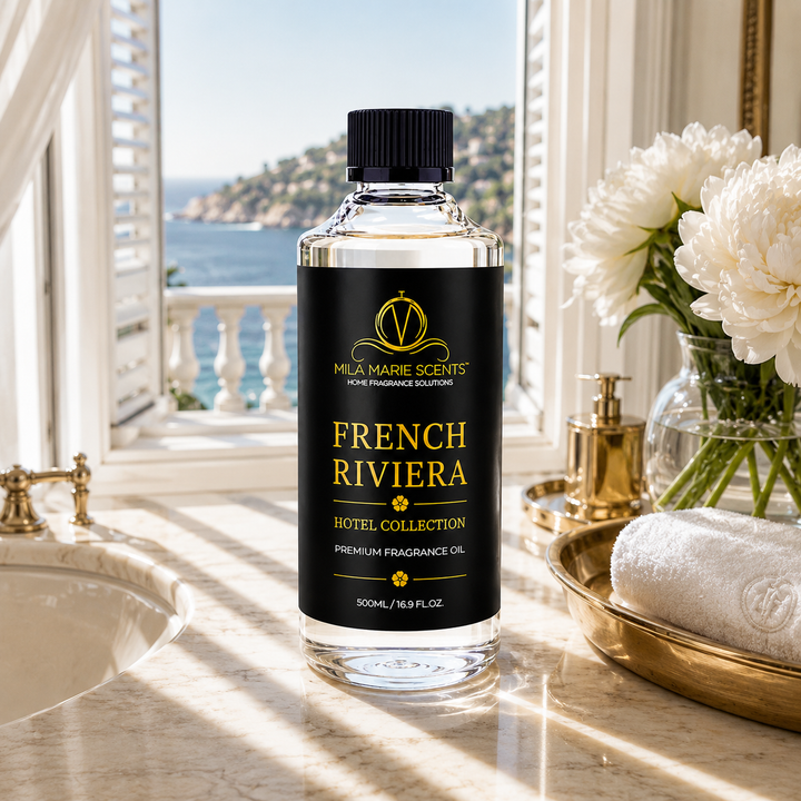 A bottle of Mla Marie Scents French Riviera Hotel Collection premium fragrance oil stands on a marble counter in a luxurious bathroom setting with a window showing a sea view, white flowers, and gold fixtures.