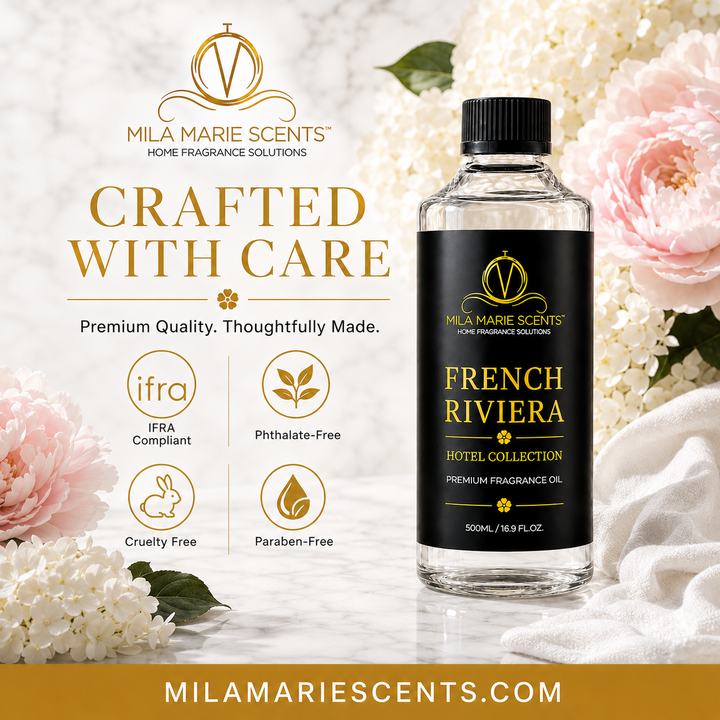 A bottle of Mila Marie Scents French Riviera Hotel Collection fragrance oil stands on a white marble surface surrounded by pink and white flowers. The bottle has a black cap and a black label with gold lettering.
