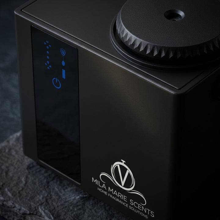 Close-up of a black portable home fragrance device with a textured top, a dark side panel showing blue indicator lights, and the white Mila Marie Scents logo on a dark textured surface.